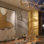 Intricate suspended sculpture hanging above dining table, with artwork on wall and lighting from spotlights