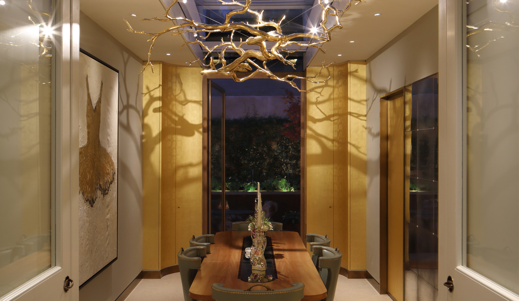 View into dining room with gold hanging tree sculpture above table with spotlights lighting from above