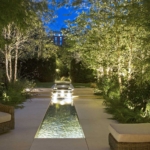 Long garden with lit water rill and uplit trees either side