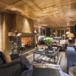 Living area of cosy chalet with wooden panelled ceiling and walls and sofas around a fireplace.